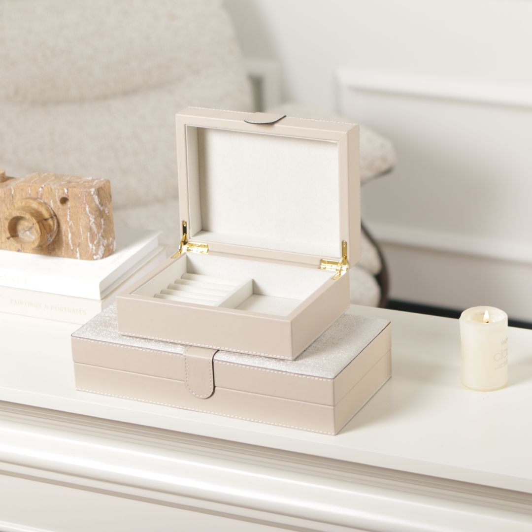 Beige hinged leather jewelry box with decorative stitching, featuring a compact rectangular design and interior compartments for jewelry storage.